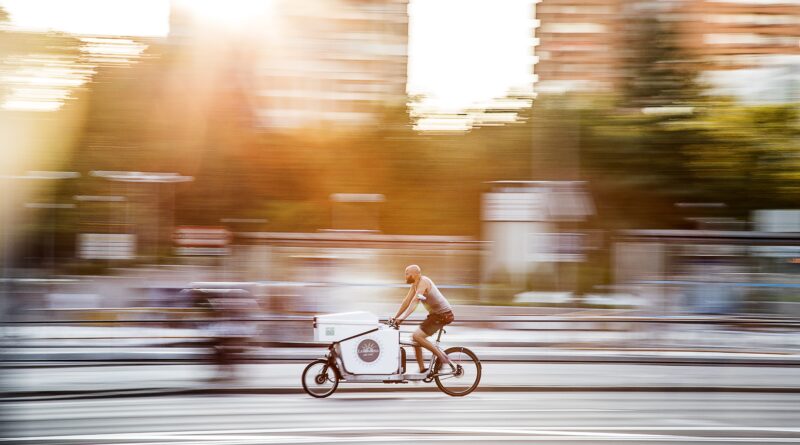 A person rides a branded cargo bicycle along a city street.